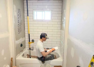 Man sits in bathtub as he works to install subway tiles to walls